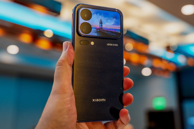 A hand holds a dark blue smartphone with a speckled finish, featuring a rectangular camera module displaying a lighthouse image and the Leica logo on the back.