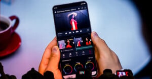 A person holds a smartphone displaying a video editing application with color grading wheels and a timeline featuring a woman in a red dress.
