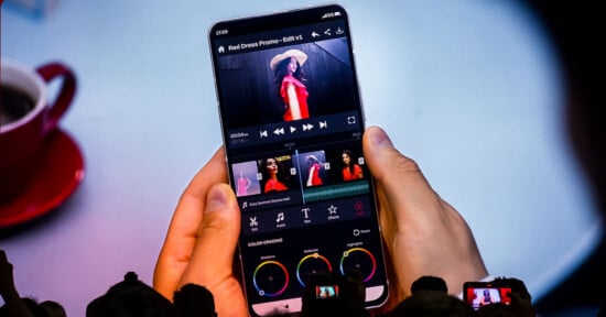 A person holds a smartphone displaying a video editing application with color grading wheels and a timeline featuring a woman in a red dress.