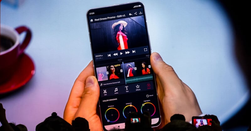 A person holds a smartphone displaying a video editing application with color grading wheels and a timeline featuring a woman in a red dress.