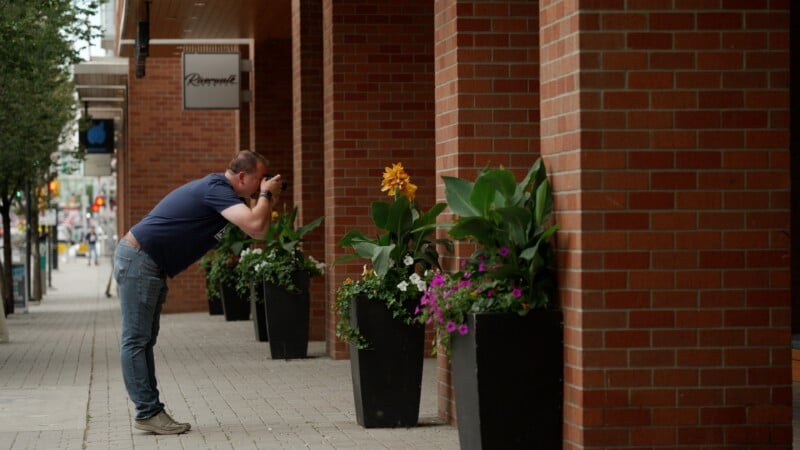 A man in a blue t-shirt and jeans leans over to photograph flowers in a black planter along a brick building sidewalk.