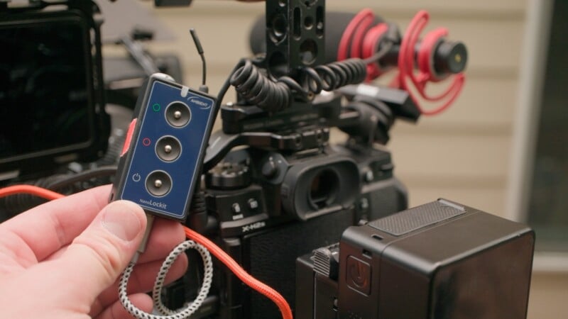 A hand holds an Ambient NanoLockit timecode generator connected by cables to a Fujifilm X-H2s camera rig.