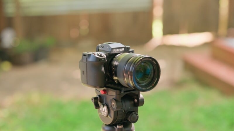 A black Fujifilm camera mounted on a tripod head with a blurred outdoor background.