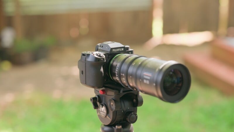 A Fujifilm camera with a zoom lens mounted on a Magnus tripod head, positioned outdoors against a blurred background.