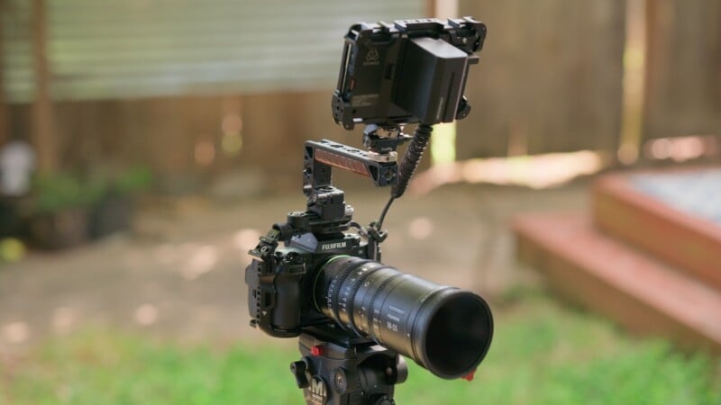 A Fujifilm camera mounted on a tripod, equipped with a zoom lens, a top handle, and an external monitor attached to the top.