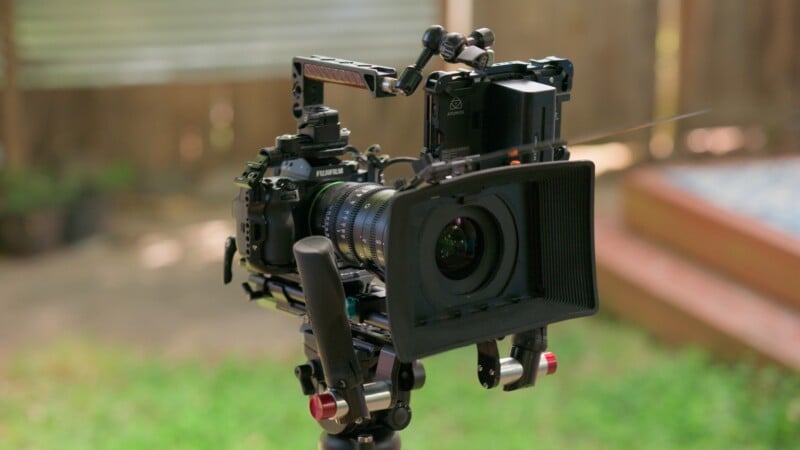 A professional cinema camera rig mounted on a tripod, featuring a lens hood, a top handle, and an external monitor attached to the back.