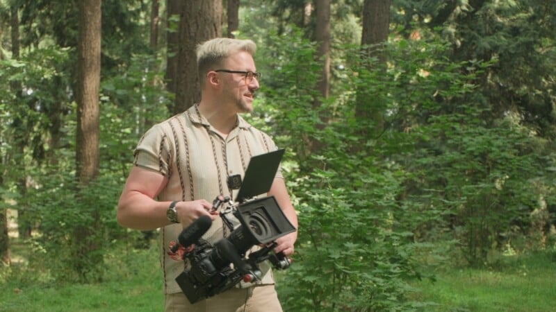 A man with blonde hair and glasses stands in a forest, holding a professional video camera with a microphone and matte box attached.