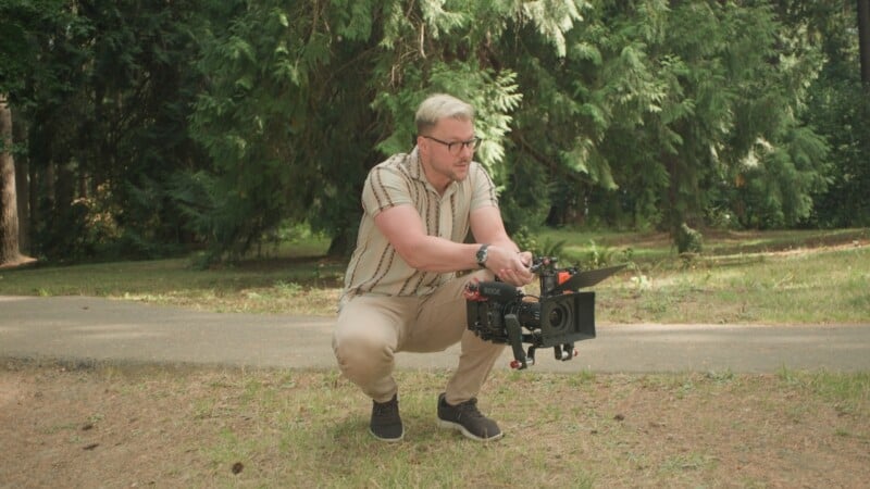 A man with blonde hair and glasses crouches in a park while holding a professional video camera rig.