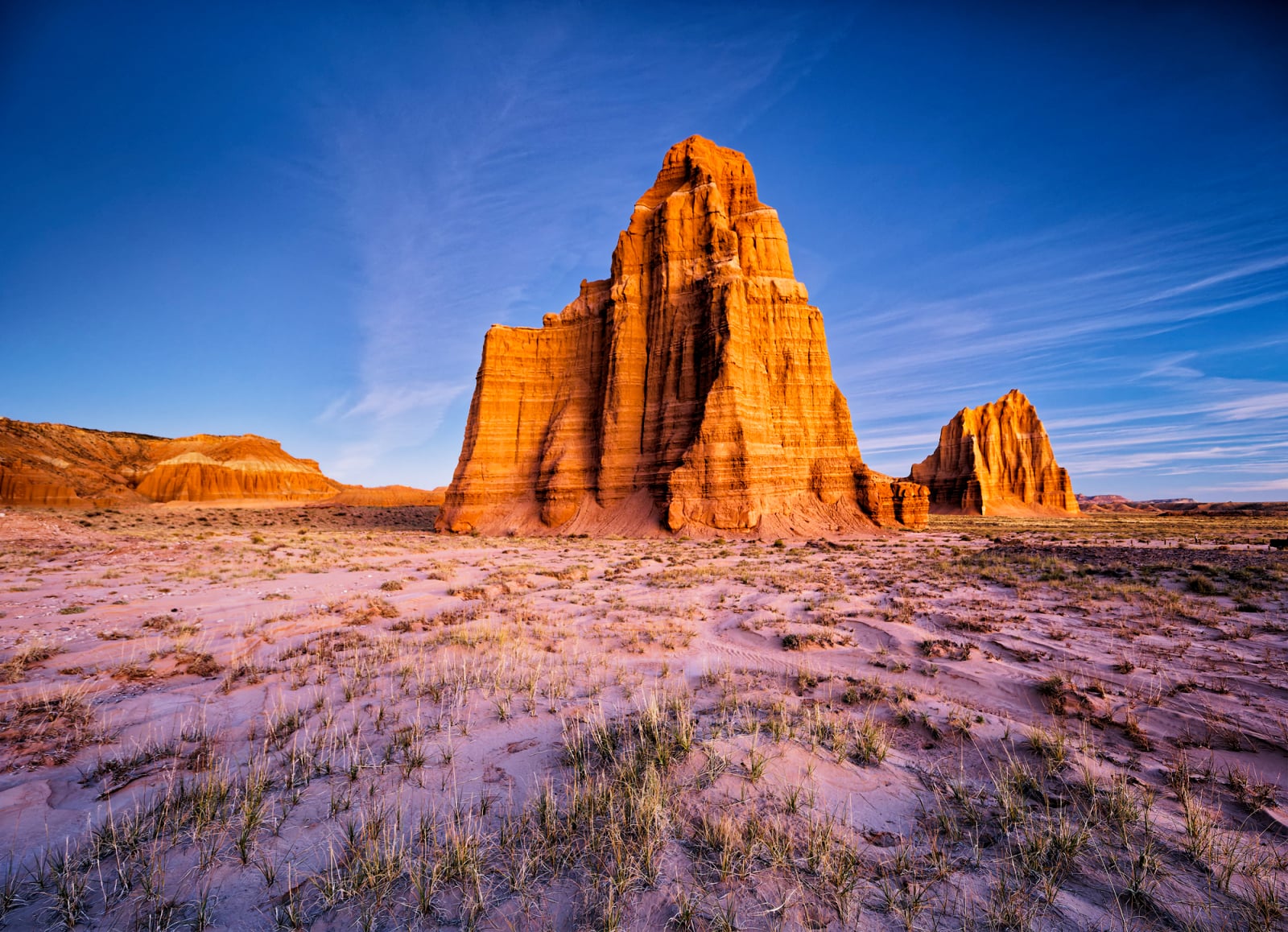 Two large, golden-orange sandstone monoliths rise from a sparse, sandy desert landscape under a clear blue sky.