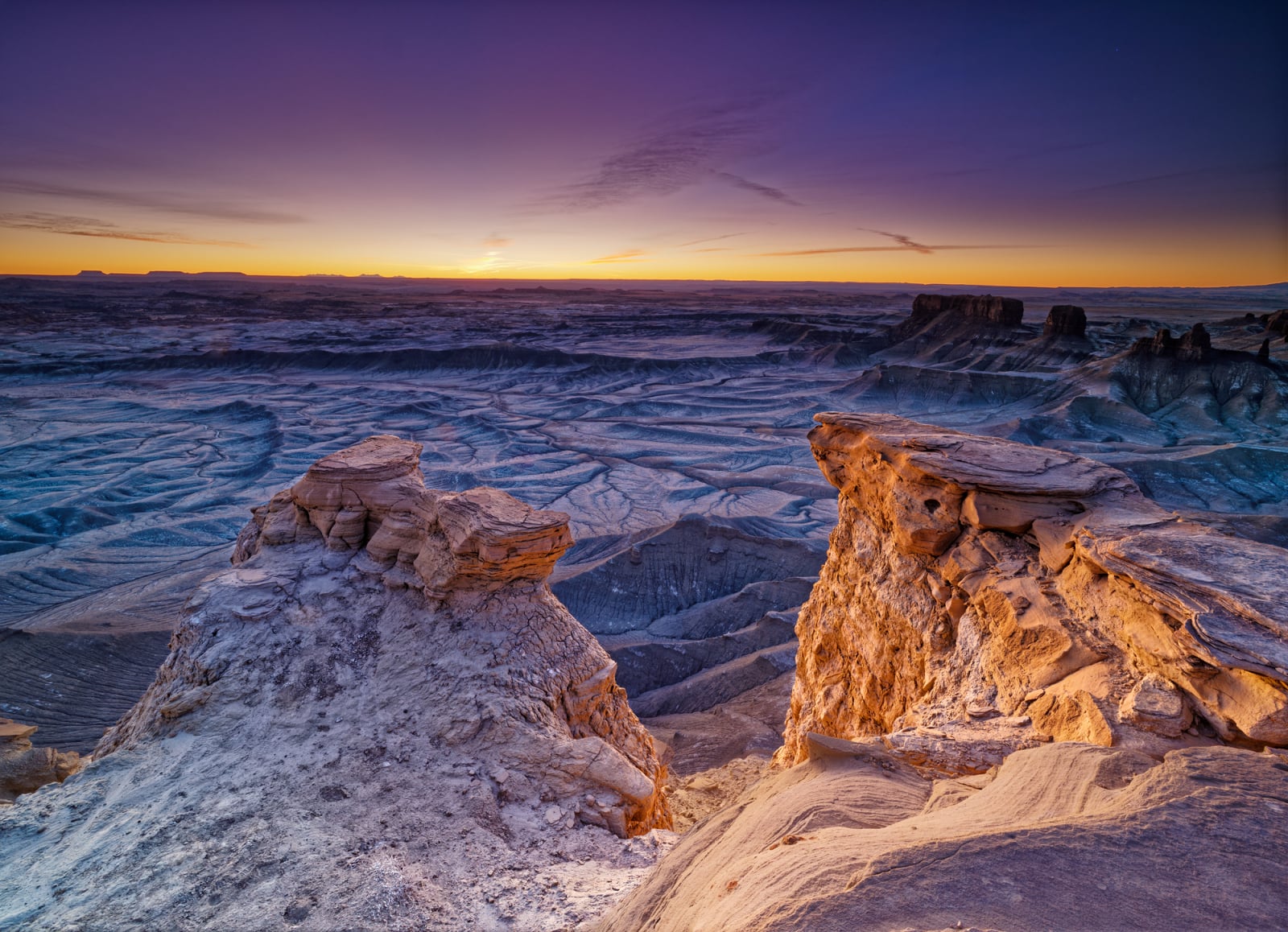 A high-angle view of a vast, rugged desert landscape at sunset, featuring prominent sandstone rock formations in the foreground.