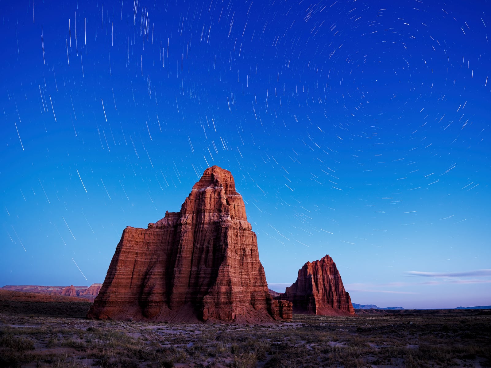 Two large, red sandstone rock formations stand in a desert landscape under a night sky with circular star trails.