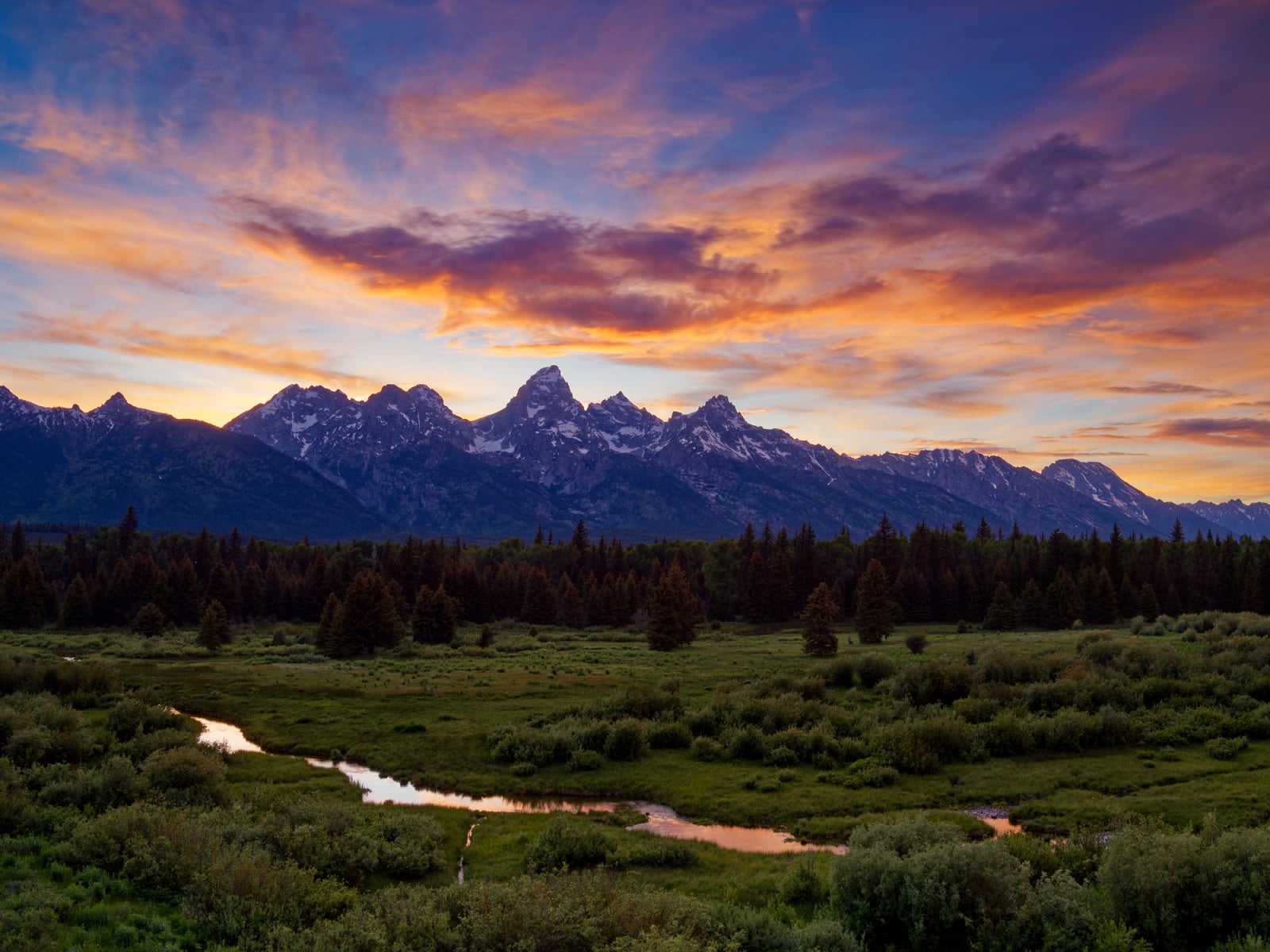 A winding stream flows through a grassy meadow toward a dense forest beneath a jagged mountain range at sunset.