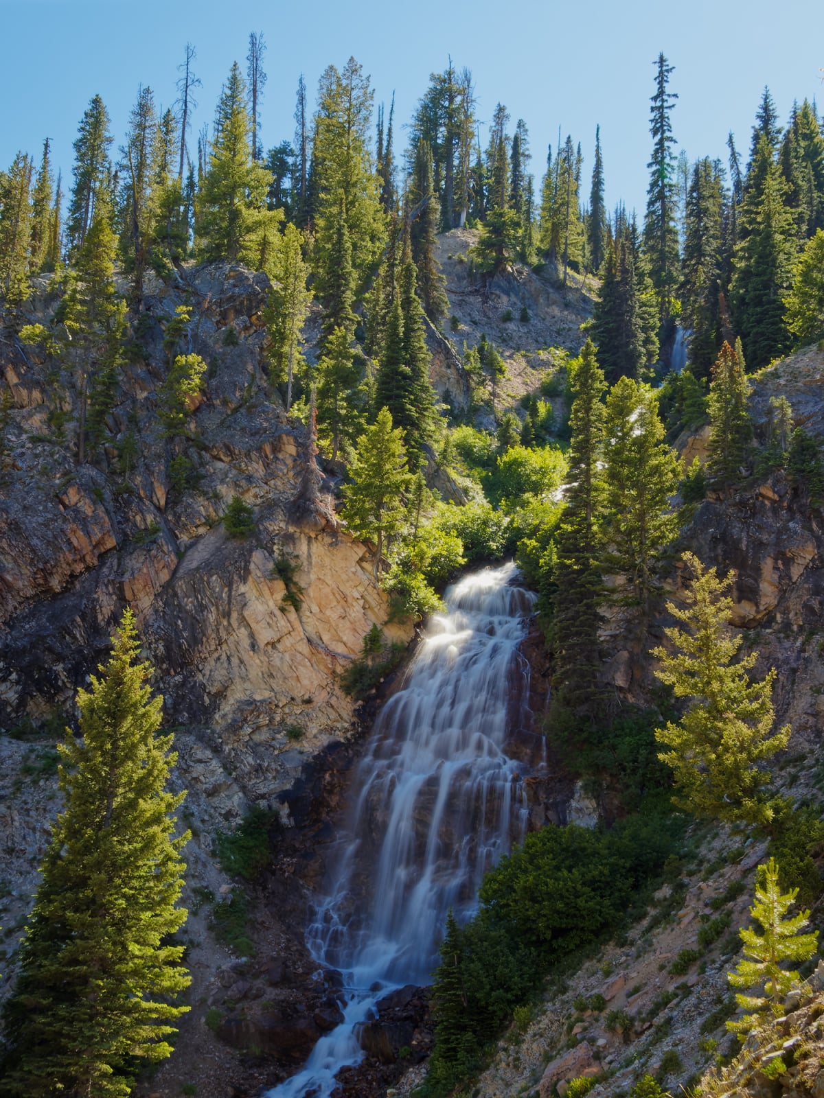 A waterfall cascades down a rocky, pine-covered mountain slope under a clear blue sky.