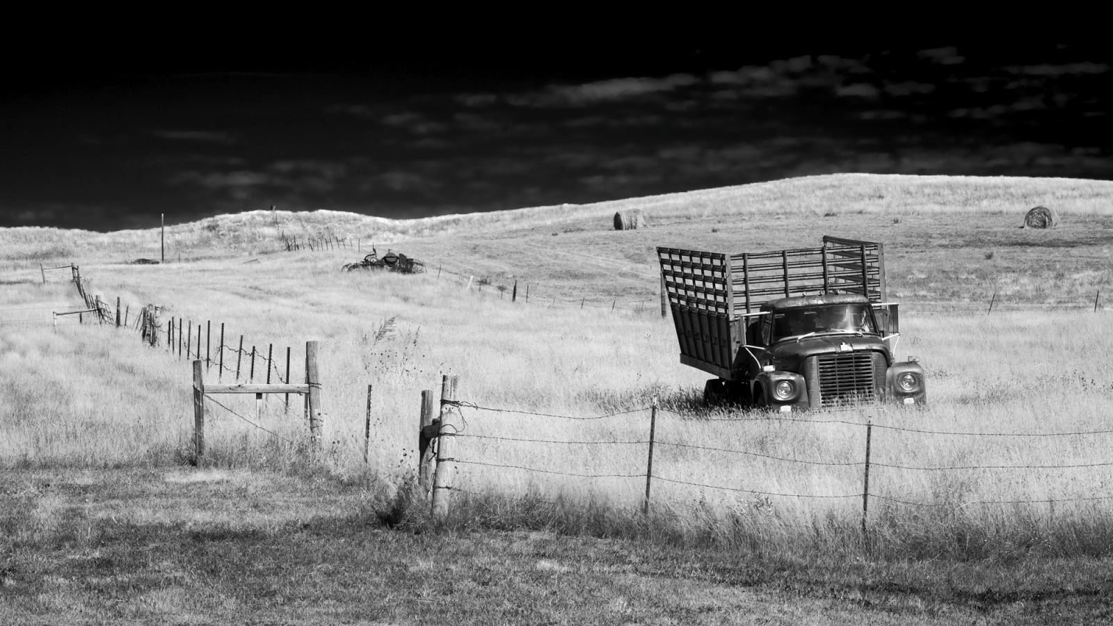 A vintage truck with a tall wooden stake bed sits in a grassy field behind a barbed wire fence, with hay bales visible on the rolling hills in the background.