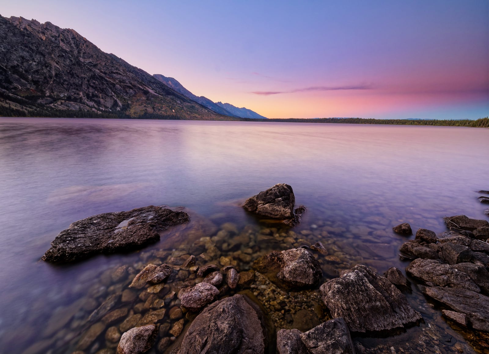 A calm lake at sunset with rocky shores in the foreground and mountains in the distance under a purple and orange sky.