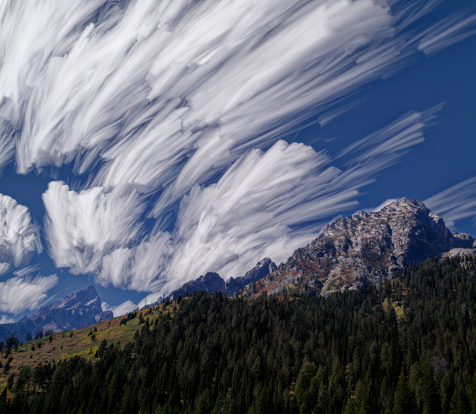 A mountain range covered in dense evergreen forests sits beneath a dramatic sky filled with long, streaking white clouds.