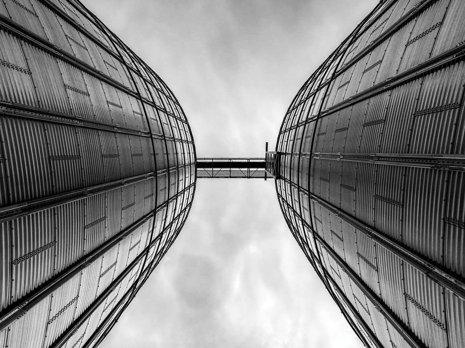 A low-angle, black-and-white view of two large, corrugated metal grain silos connected by a narrow bridge against a cloudy sky.