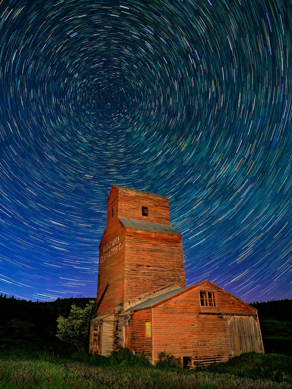 A weathered wooden grain elevator stands in a field under a night sky filled with circular star trails.