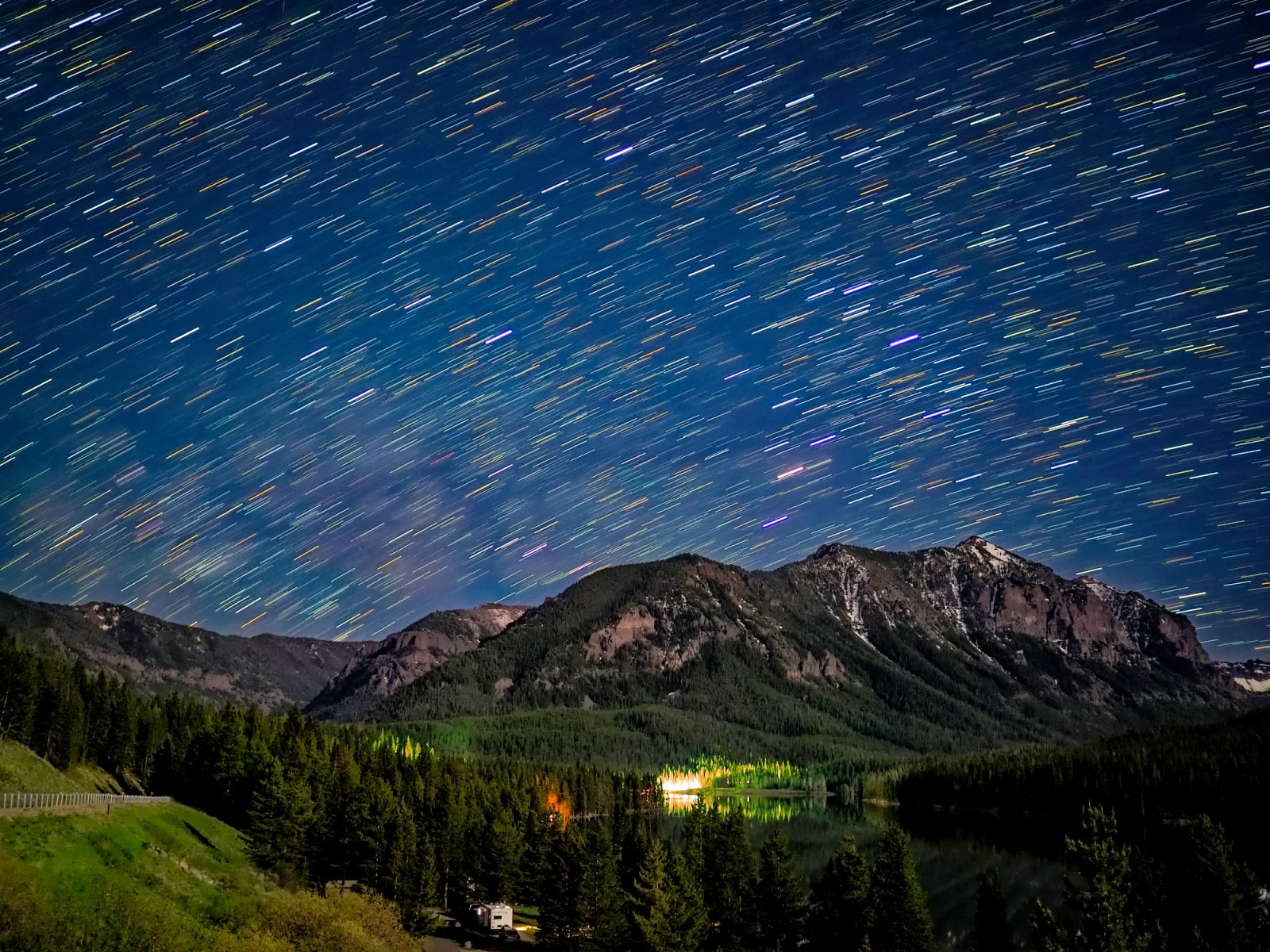 A long-exposure night shot shows star trails streaking across a dark blue sky above a mountain range and a forested lake.