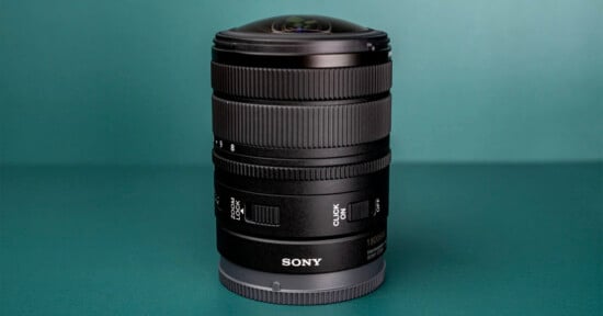 A black Sony camera lens stands upright on a teal surface, featuring a prominent glass element at the top and various control rings and switches on its barrel.