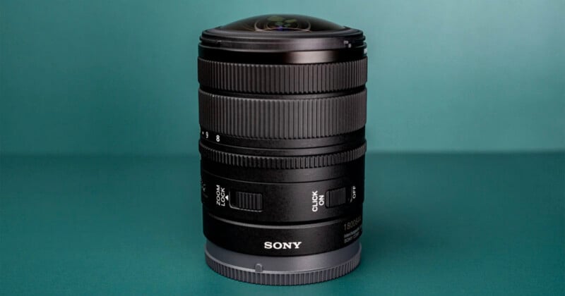 A black Sony camera lens stands upright on a teal surface, featuring a prominent glass element at the top and various control rings and switches on its barrel.