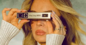 A person with blonde hair holds a silver Camp Snap 110 D camera in front of their eyes, wearing a white ribbed sweater.