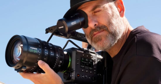 A man with a beard and a black hat looks through the viewfinder of a professional Fujifilm cinema camera equipped with a Fujinon 32-90 lens.