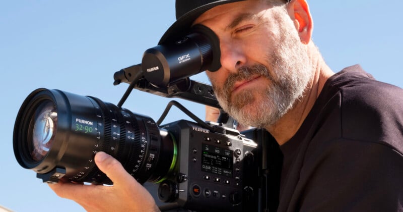 A man with a beard and a black hat looks through the viewfinder of a professional Fujifilm cinema camera equipped with a Fujinon 32-90 lens.