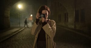 A woman in a tan trench coat stands on a cobblestone street at night, holding a camera up to her face to take a picture.