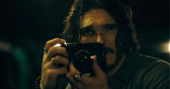 A man with long, dark, wavy hair and a mustache holds a black camera up to his face to take a photograph.