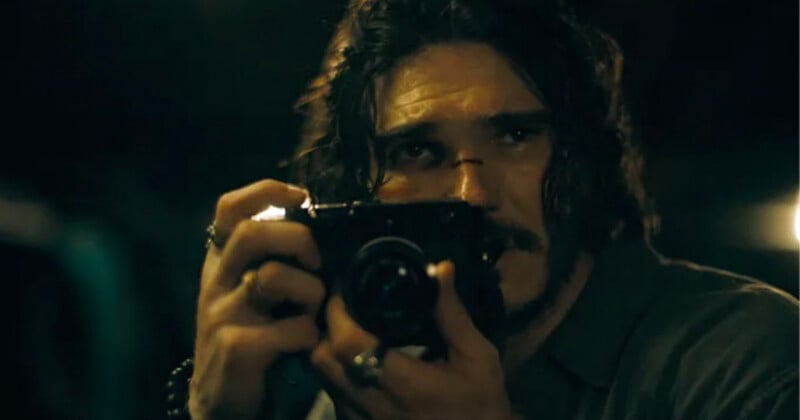 A man with long, dark, wavy hair and a mustache holds a black camera up to his face to take a photograph.