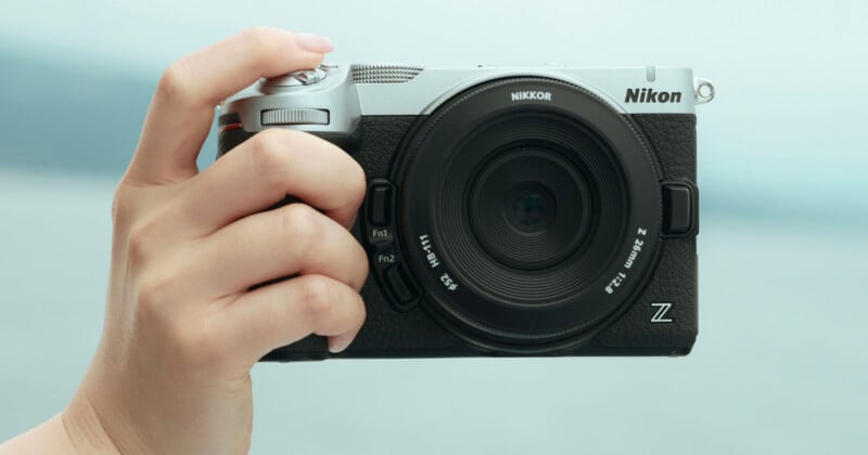 A hand holds a silver and black Nikon Z mirrorless camera with a Nikkor Z 26mm f/2.8 lens against a blurred light blue background.