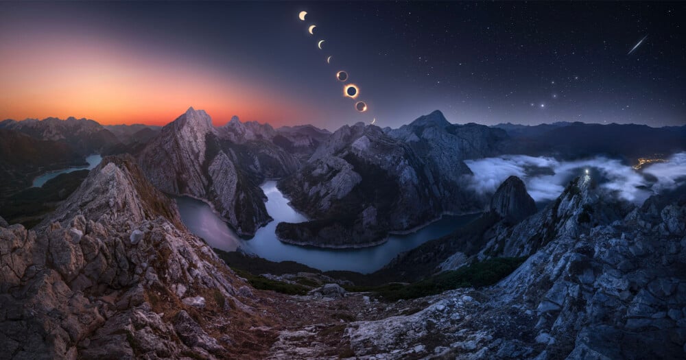 A composite view of a solar eclipse over a winding river valley surrounded by jagged, rocky mountains at twilight.