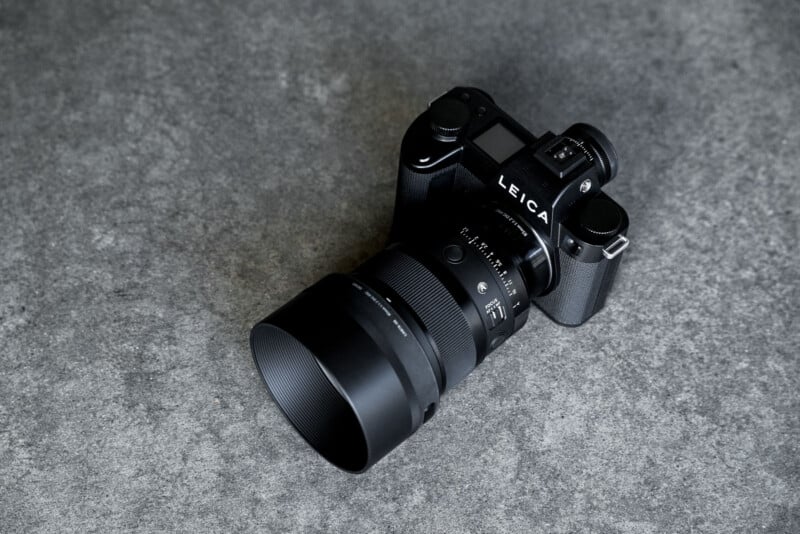 A black Leica mirrorless camera with a large Sigma 85mm lens attached, resting on a textured gray concrete surface.