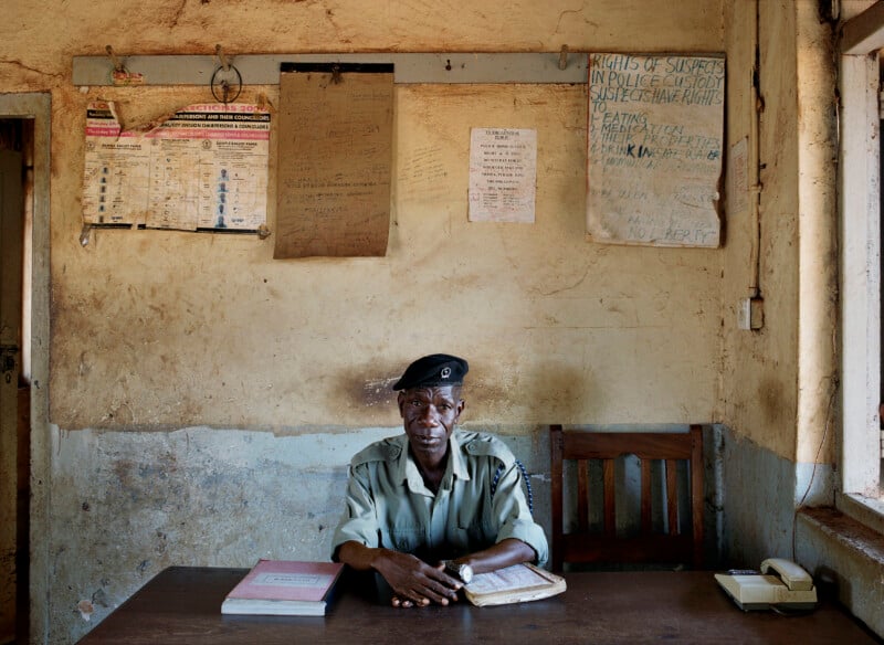 A police officer in a uniform and beret sits at a wooden desk in a sparse office with handwritten signs and election posters on the wall.