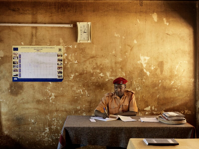 A person in a tan uniform and red beret sits at a desk writing in a notebook against a weathered wall with a calendar and clock.