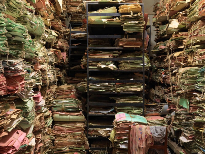 A dense, floor-to-ceiling archive of paper files and documents stacked on metal shelving units and piled directly on the floor.