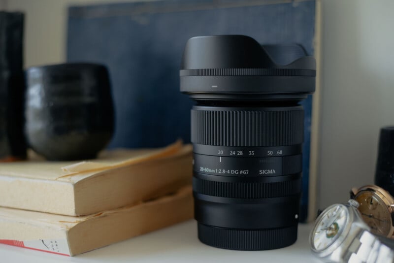 A Sigma 20-60mm lens stands upright on a white surface next to a stack of books, a dark ceramic cup, and two wristwatches.