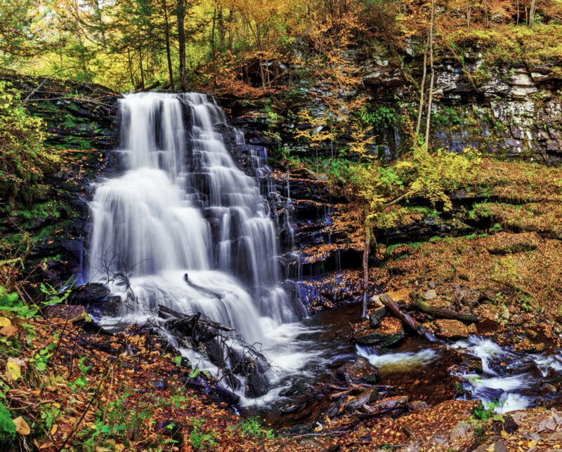 A tiered waterfall cascades over dark, mossy rocks surrounded by autumn foliage and fallen leaves in a forest.
