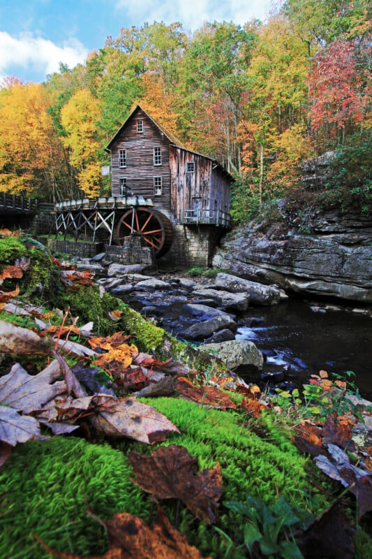 A rustic wooden grist mill with a large water wheel sits beside a rocky stream, surrounded by a forest with vibrant autumn foliage.