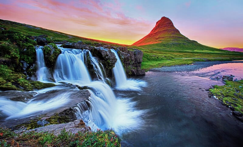 A multi-tiered waterfall flows into a stream beneath a prominent, cone-shaped mountain under a colorful sunset sky.