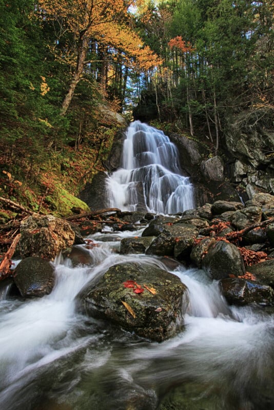 A cascading waterfall flows over dark, mossy rocks surrounded by a dense forest with autumn-colored foliage.