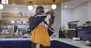 A man with braided hair wearing a black and orange jacket holds a camera up to his face to take a picture in a cafe.