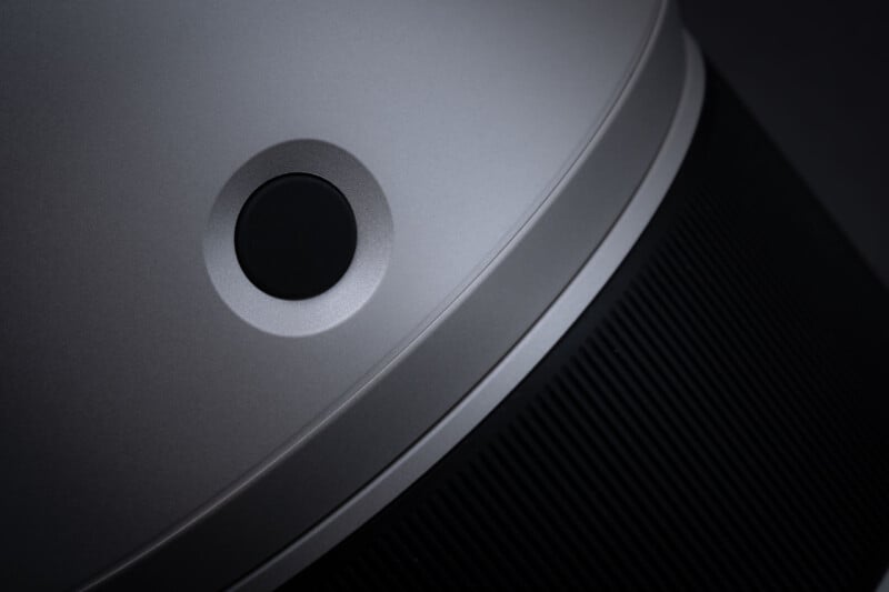 A close-up view of a matte silver surface featuring a circular black button set into a recessed, beveled area, with a black ribbed texture visible on the right side.