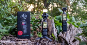A black product box labeled "Xtra Muse 2 Pro" stands next to two handheld gimbal cameras on tripods, one with a small light attached, set against a background of green foliage and a tree stump.