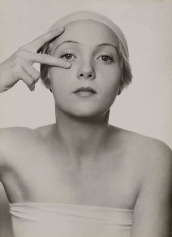 A black-and-white portrait of a woman wearing a swim cap, holding two fingers to her right eye.