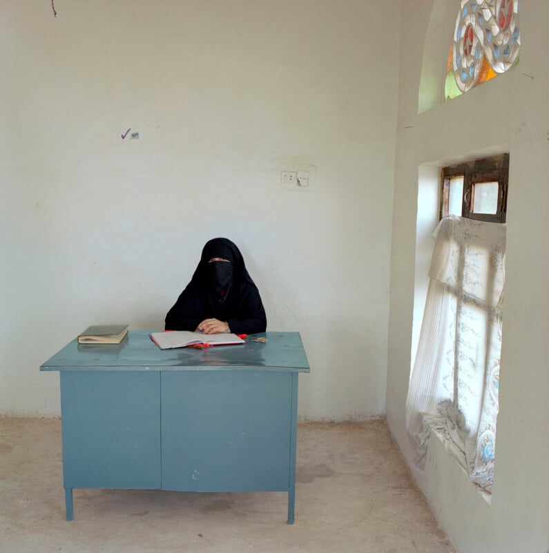 A person wearing a black niqab and abaya sits at a blue metal desk with an open book in a sparsely furnished room.