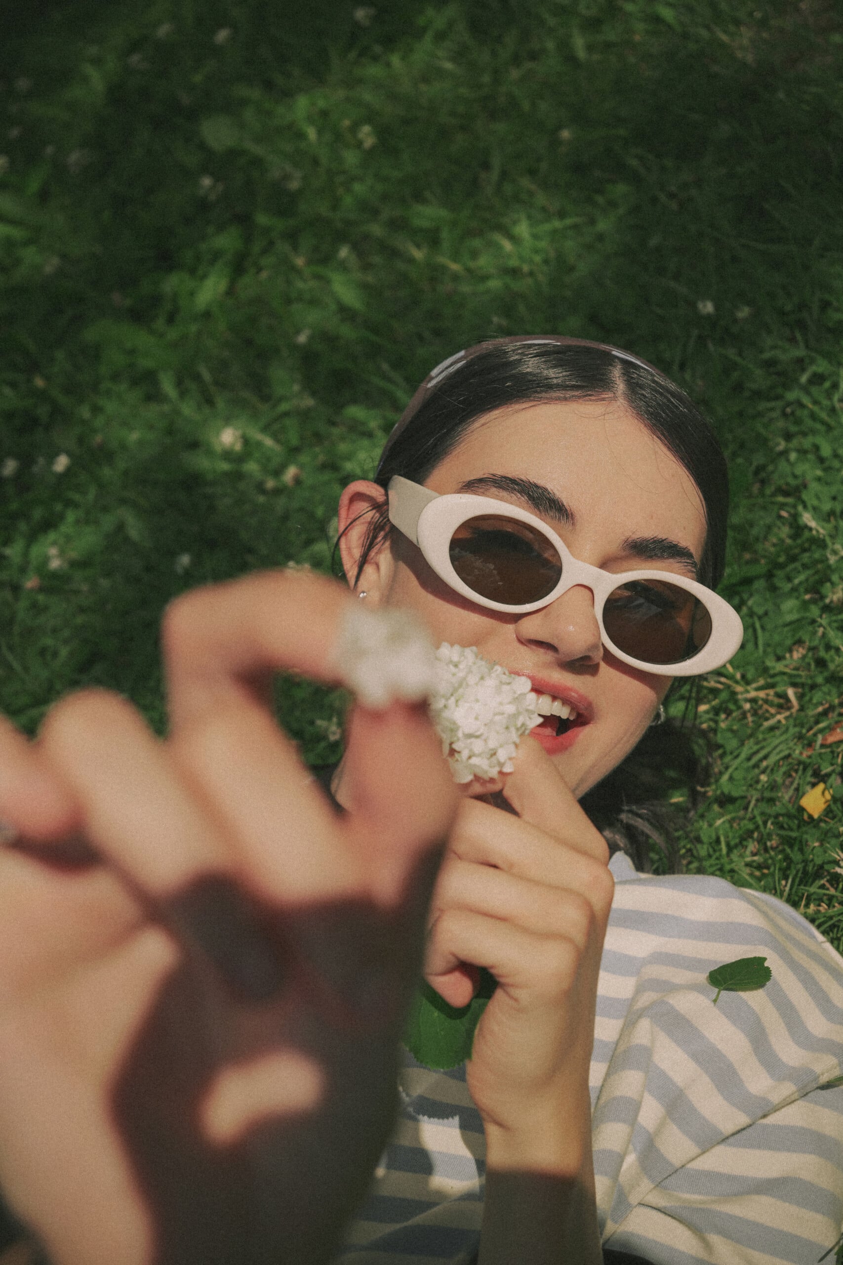 A person wearing white oval sunglasses and a striped shirt lies in the grass, holding a small white flower near their mouth.