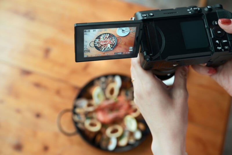 A person holds a digital camera with an articulating screen, which displays a live view of a seafood paella dish sitting on a wooden table.