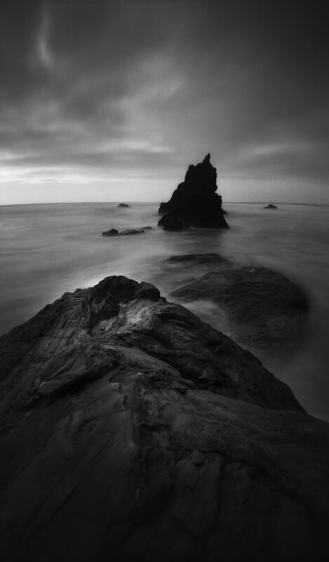 A black and white long-exposure shot of a jagged rock formation rising from a calm, misty ocean with large rocks in the foreground.